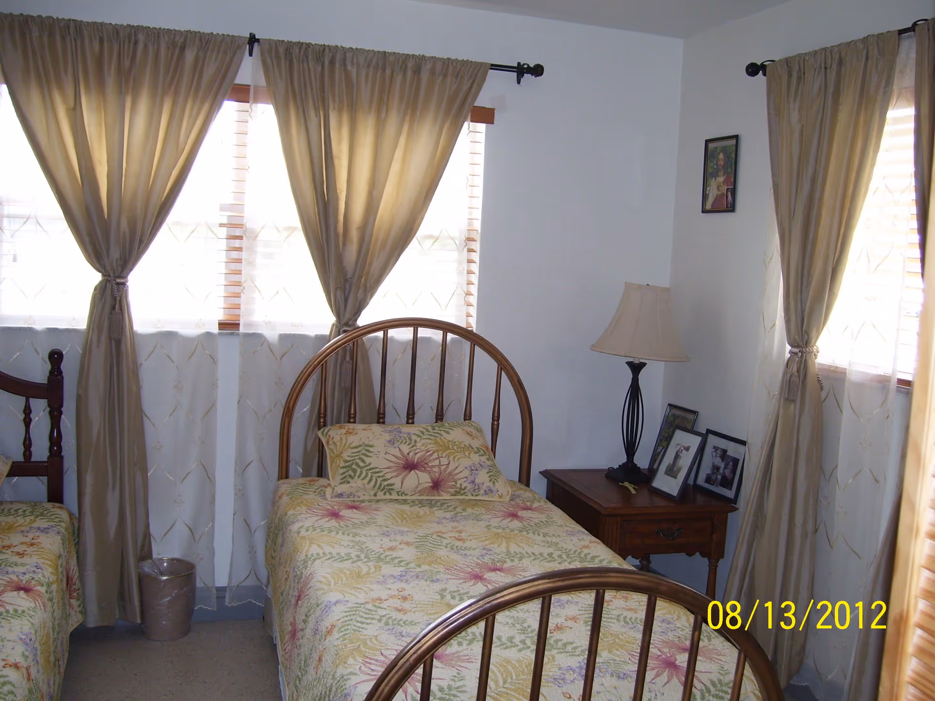 A bedroom with two single beds featuring floral bedspreads. The room has three windows with beige curtains tied back, a wooden nightstand with a lamp and framed photos, and white walls.