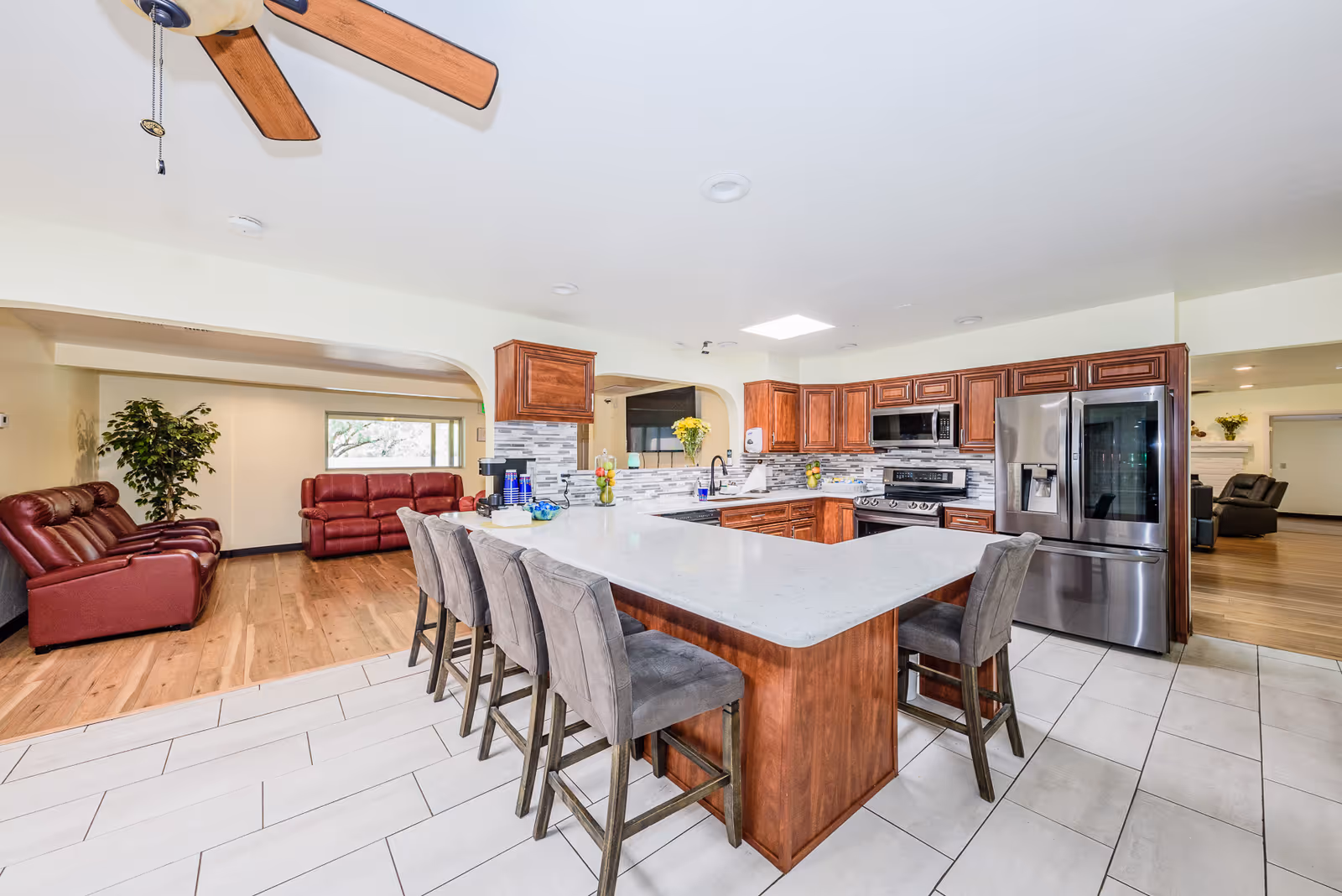 Spacious kitchen area with a large white marble island surrounded by six gray upholstered bar stools. The kitchen features wooden cabinets, stainless steel appliances including a refrigerator, oven, and microwave. Adjacent to the kitchen is a living area with red leather sofas, a large window, and a potted plant. The floor transitions from white tiles in the kitchen to wood flooring in the living area. A ceiling fan with wooden blades is visible above.