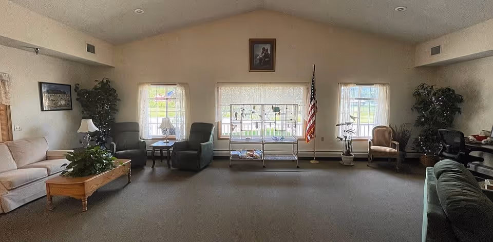 A spacious living room with beige walls and carpeted floor. The room features a beige sofa on the left, two green armchairs with a small table and lamp between them near the center windows, and a green couch on the right. There are three large windows with sheer curtains letting in natural light. An American flag stands near the right window, and there are several potted plants and framed pictures on the walls. A metal rack with various items is placed in front of the middle window.