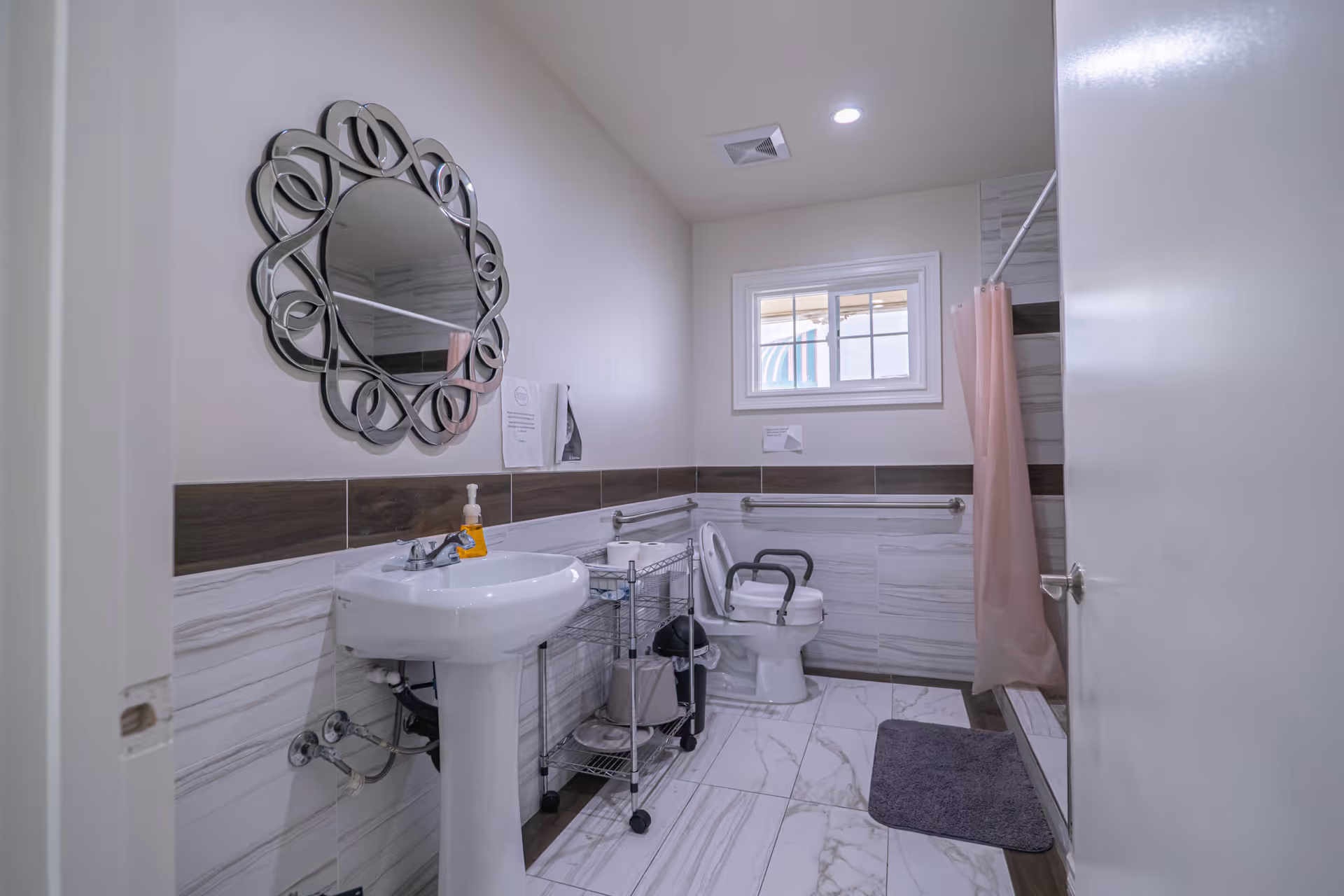 Accessible bathroom with a pedestal sink and decorative round mirror, a toilet with safety rails, a shower with a pink curtain, and tiled floors.