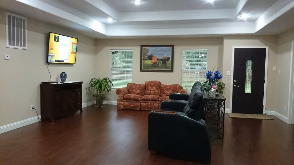 A senior living facility common area with wooden flooring, a red patterned sofa against the back wall between two windows with blinds, two black recliner chairs facing a wall-mounted TV, a dark wooden cabinet below the TV with a decorative vase on top, a small table with blue flowers and a small American flag near a dark front door with a glass panel, and a framed picture hanging on the wall above the sofa.