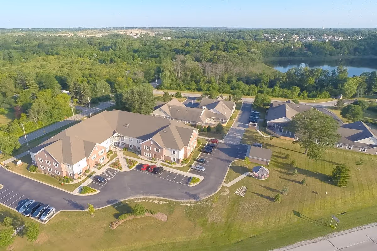 Aerial view of Frontida Assisted Living: Clifden Court facility showing multiple buildings with parking lots, surrounded by green trees and a nearby lake in the background under a clear sky.