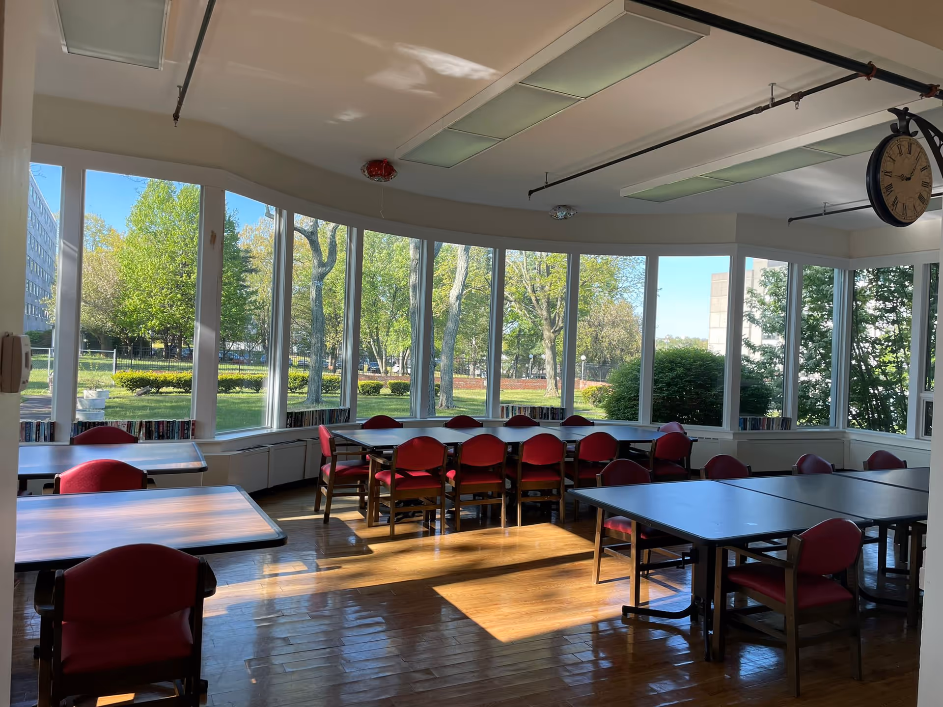 A bright room with large windows showing a green outdoor area with trees and bushes. Inside, there are several tables with red cushioned chairs arranged around them. The floor is wooden, and there is a large clock hanging from the ceiling.