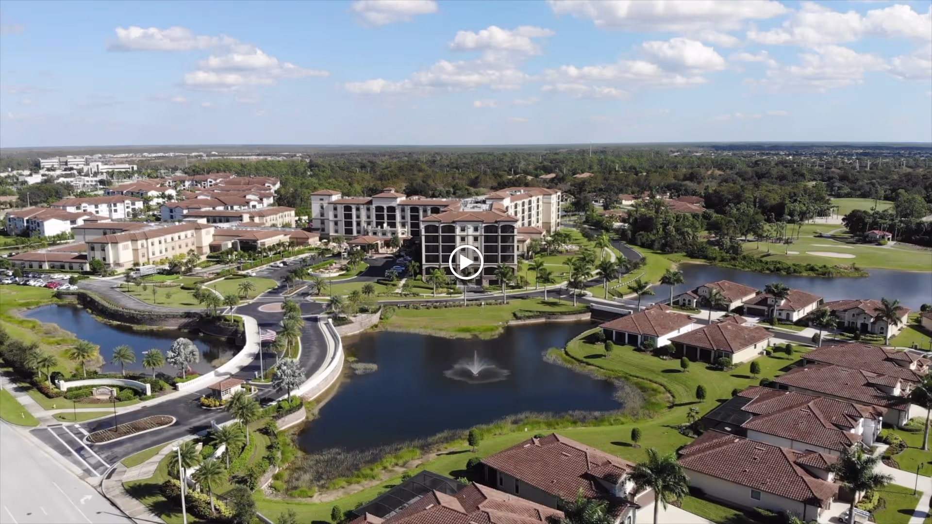 Aerial view of The Arlington of Naples senior living facility showing multiple buildings with red-tiled roofs, a central pond with a fountain, surrounding greenery, palm trees, and a clear blue sky with scattered clouds.