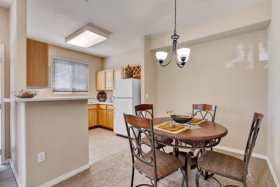 A dining area with a round wooden table and four chairs with patterned cushions. The table is set with placemats and a decorative centerpiece. Adjacent to the dining area is a kitchen with light wood cabinets, a white refrigerator, a window with blinds, and a countertop with a fruit bowl.