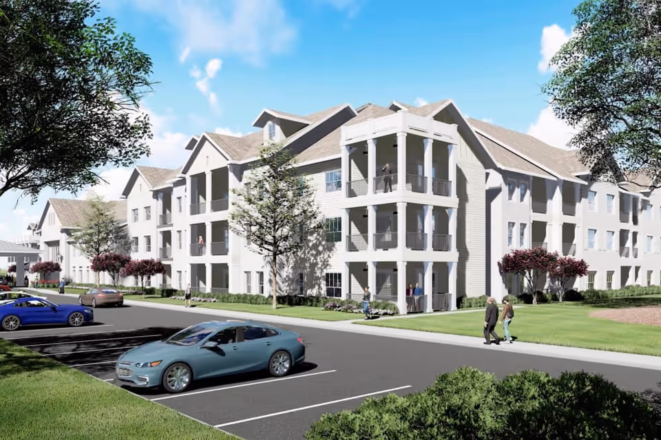 Exterior view of a multi-story senior living facility building with balconies, surrounded by trees, grass, and a parking lot with several cars. People are walking and standing near the building under a clear blue sky.