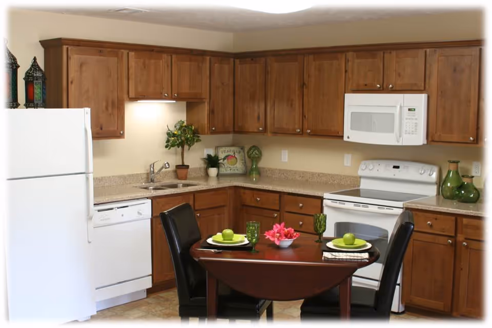 A kitchen with wooden cabinets, a white refrigerator, dishwasher, stove, and microwave. There is a small dining table with two black chairs, set with green plates, green glasses, and a centerpiece of pink flowers. Decorative items are placed on the countertops and above the refrigerator.
