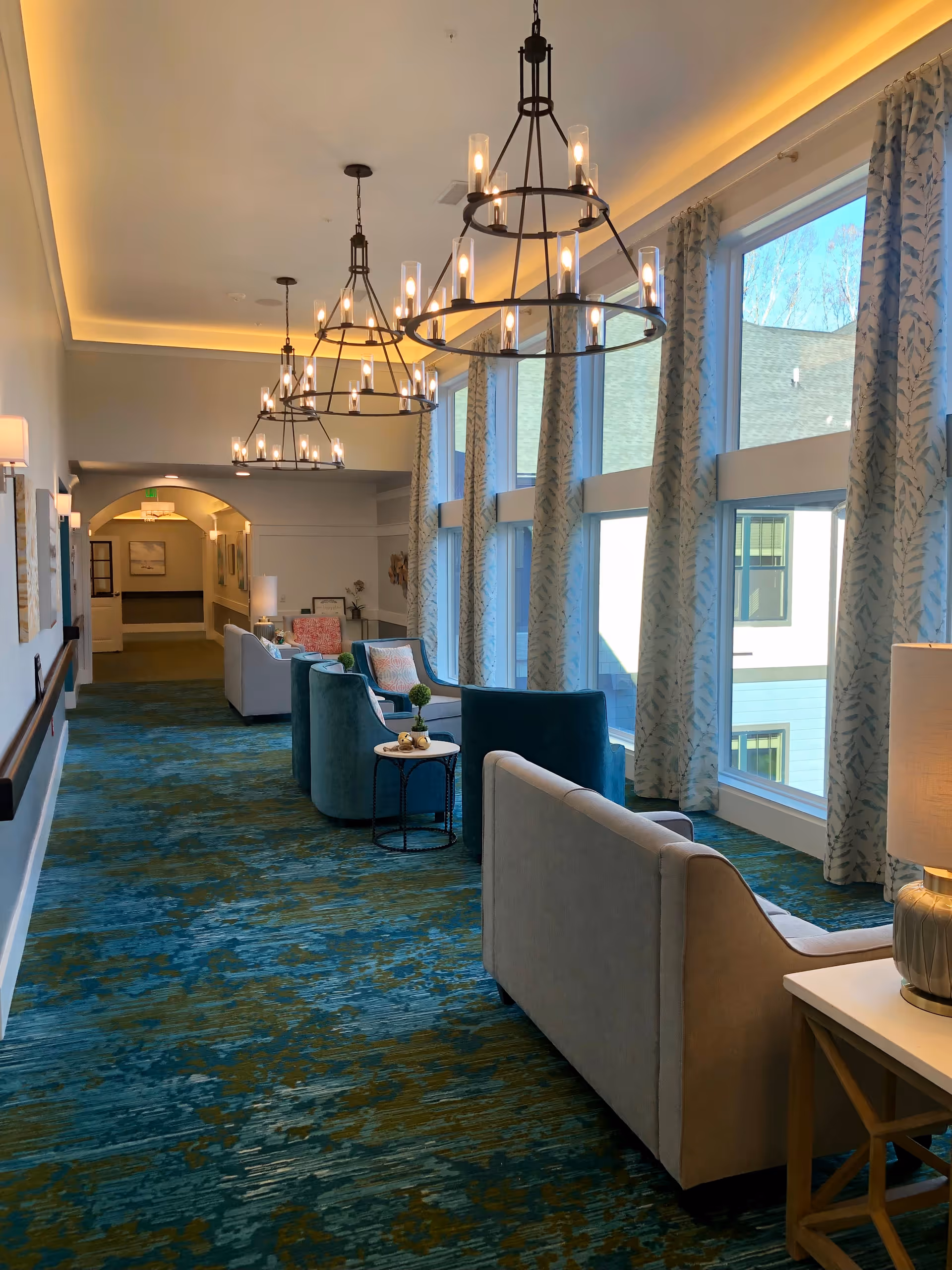 A bright and spacious hallway in a senior living facility with large windows on one side covered with patterned curtains. The hallway is furnished with comfortable armchairs and sofas arranged in seating areas along the windows. Modern chandeliers hang from the ceiling, and the carpet has a blue and green abstract pattern. A side table with a lamp is visible in the foreground.