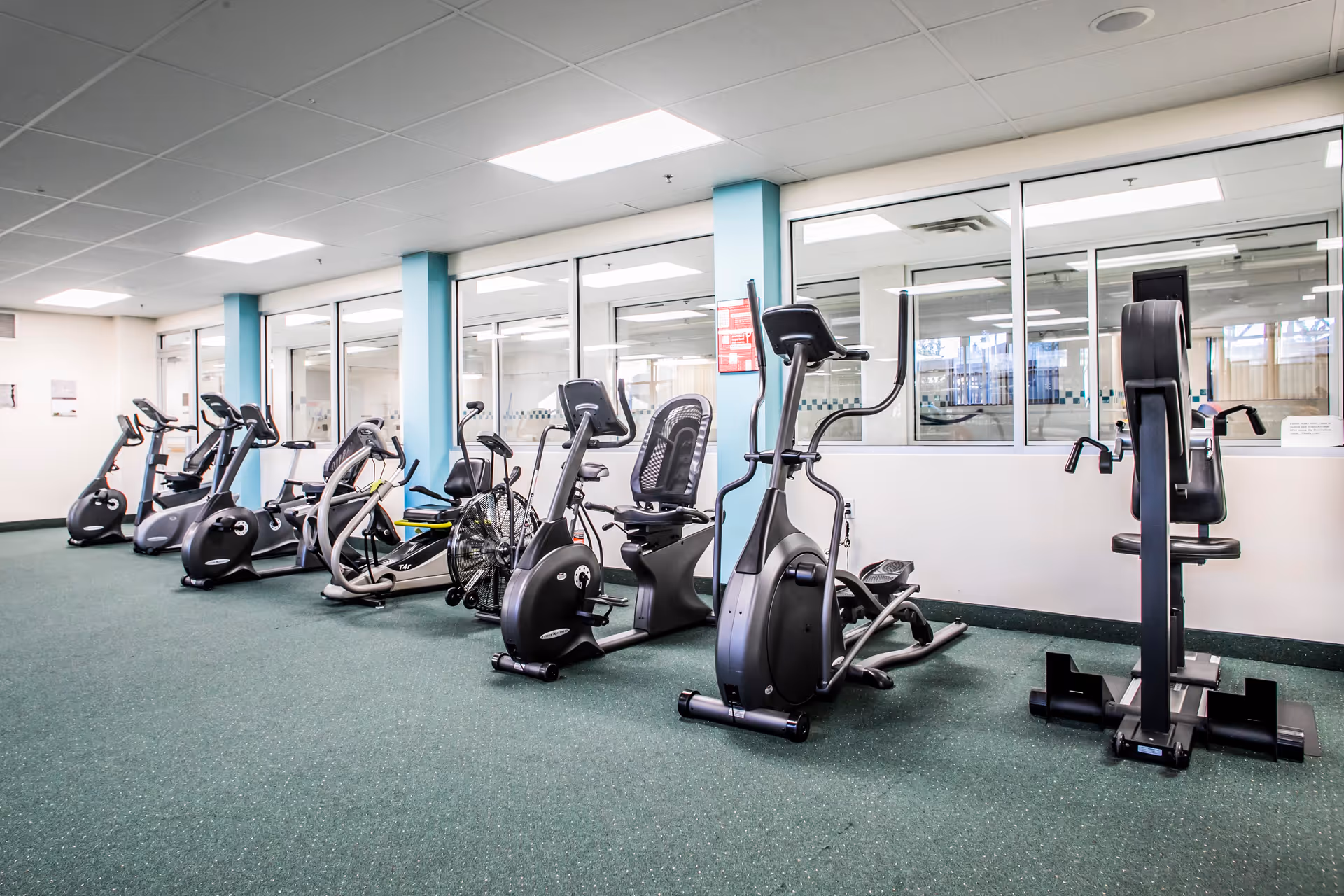 A fitness room with various exercise equipment including stationary bikes, an elliptical machine, and a rowing machine. The room has green carpet flooring, white walls with blue pillars, and large windows looking into another room.