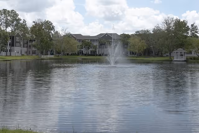 View of a senior living building across a pond with a central fountain and a gazebo on a cloudy day.