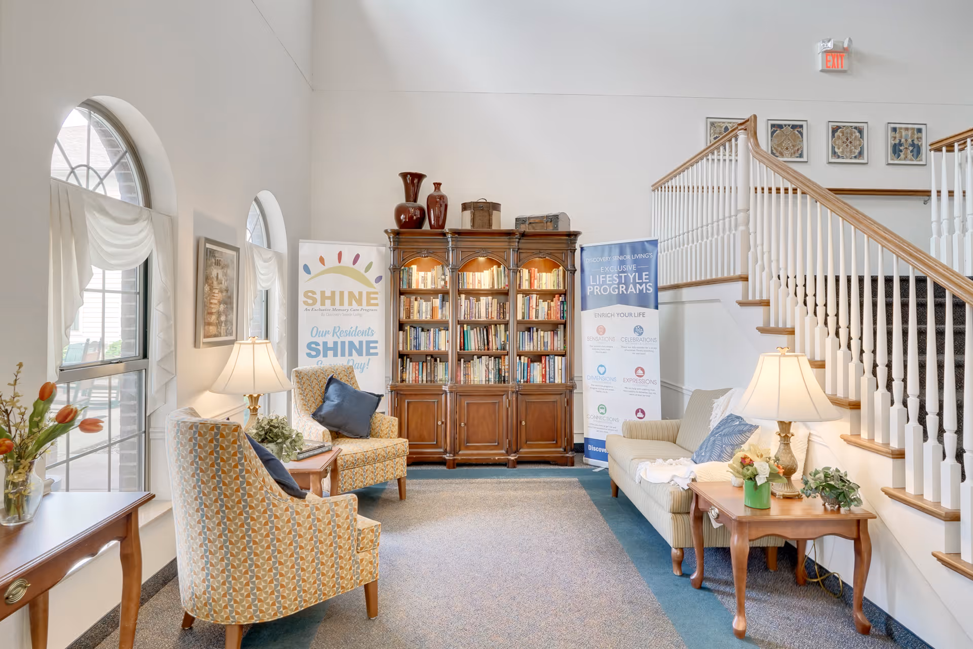 A cozy living room area in a senior living facility with two patterned armchairs, a beige sofa, wooden side tables with lamps, a large wooden bookshelf filled with books, and two banners promoting lifestyle programs. The room has arched windows with white curtains and a staircase with white railings and wooden handrails.