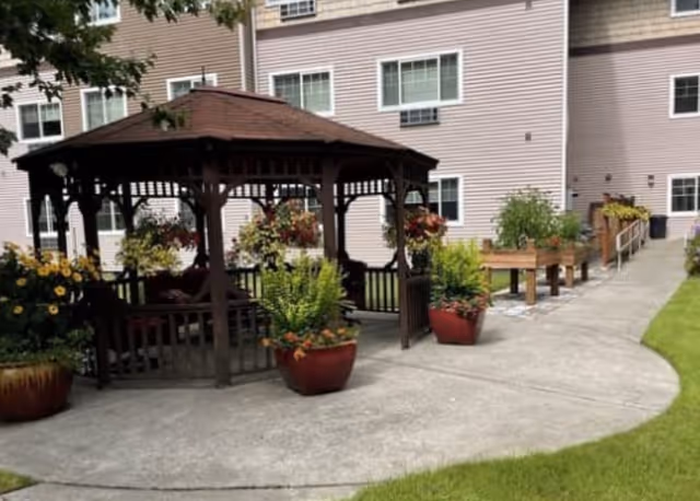 A wooden gazebo surrounded by potted plants and raised planters on a concrete courtyard outside a multi-story residential building.