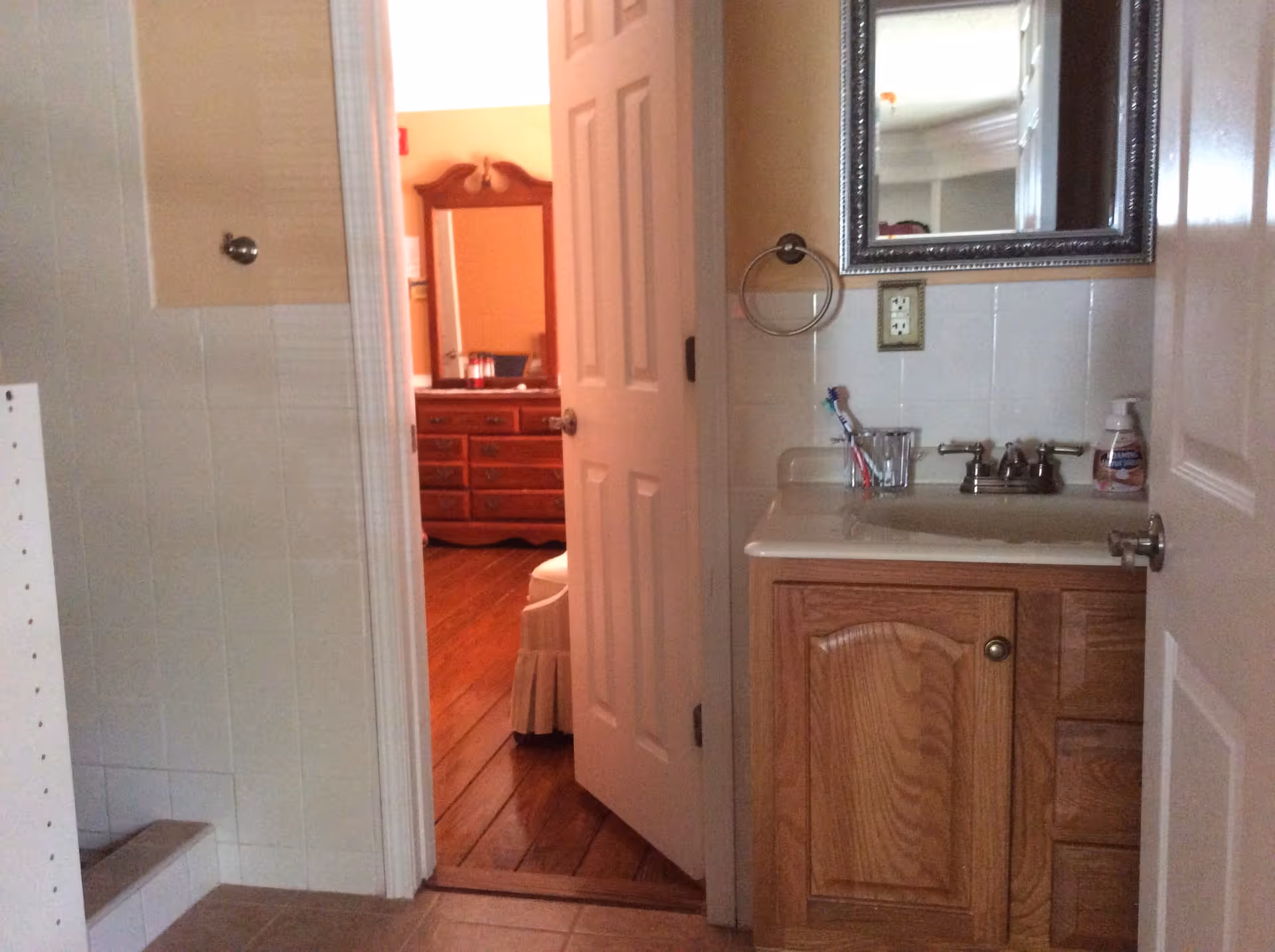 View of a bathroom with a wooden vanity cabinet, a sink with a faucet, a mirror above the sink, a toothbrush holder with toothbrushes, and a soap dispenser. The bathroom door is open, revealing a glimpse of a bedroom with wooden flooring, a dresser with a mirror, and part of a bed with a skirted bedspread.