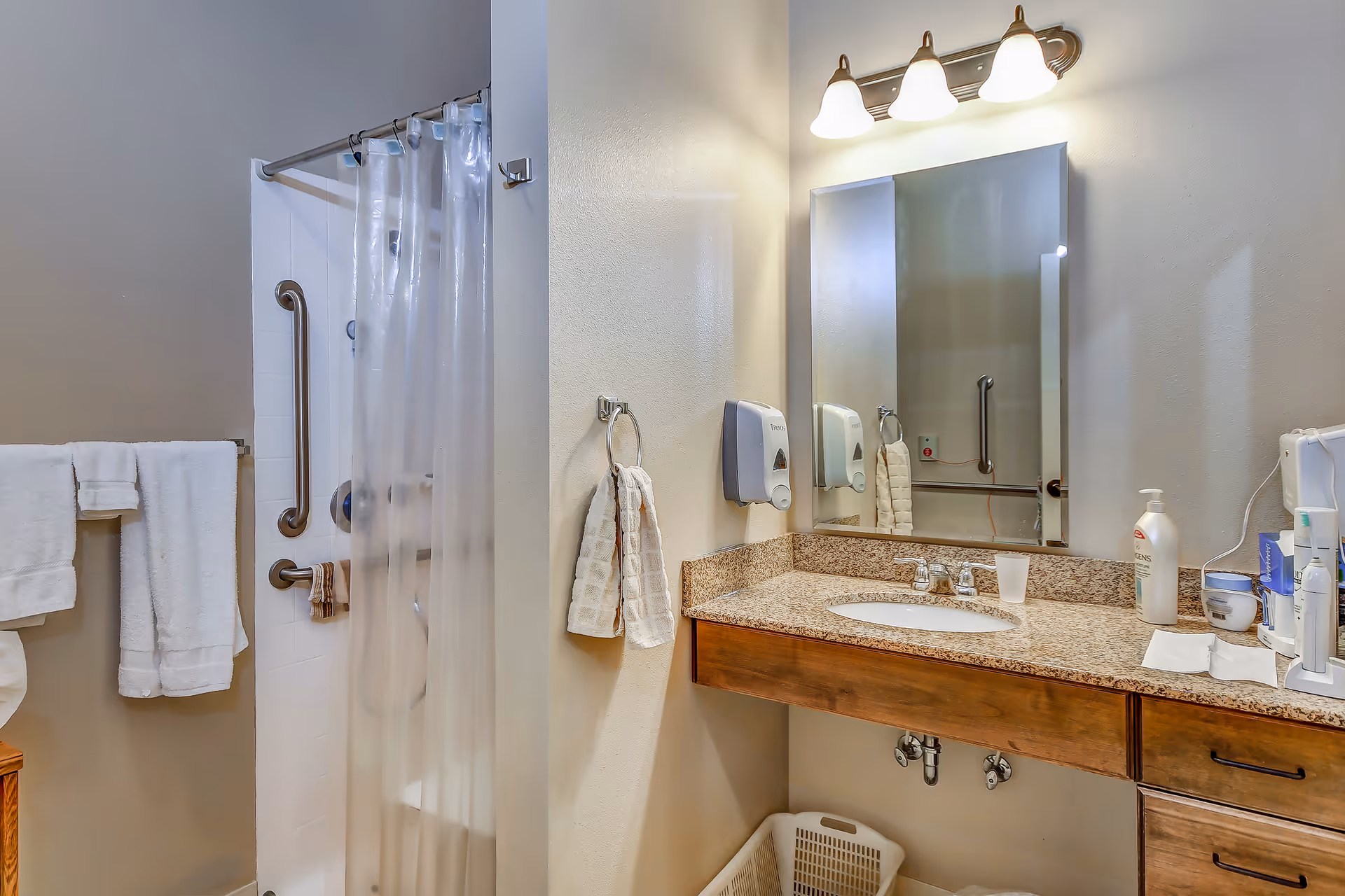 A bathroom with a shower area featuring a clear plastic curtain and grab bars, a granite countertop with a sink, a large mirror above the sink, and various toiletries on the counter. There are white towels hanging on a towel rack and a hand towel on a ring near the sink. The lighting fixture above the mirror has three lights.