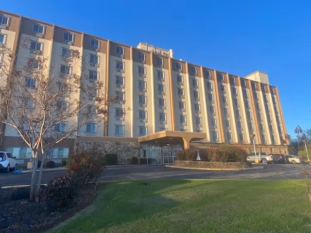 Exterior view of a multi-story senior living facility building under a clear blue sky, with a driveway and landscaped lawn in front.