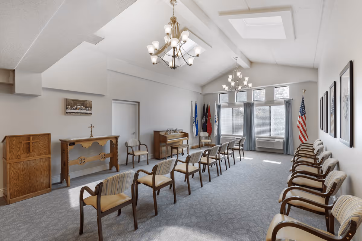 A bright, spacious room with rows of wooden chairs arranged facing a small altar and a piano. The room has high ceilings with two chandeliers, large windows with blue curtains, and several flags including an American flag. The walls are decorated with framed pictures.