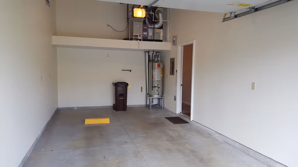Empty attached garage with concrete floor, a water heater and HVAC unit at the back, a trash bin, and an open door to the interior.