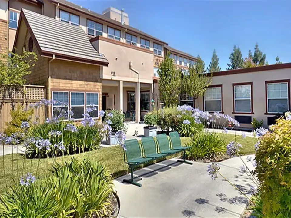 Courtyard with a green three-seat bench, flowering beds, and the exterior of a multi-story assisted living building.
