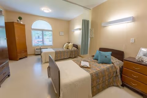 A bright nursing facility bedroom with two single beds, dressers, a privacy curtain, and a large arched window.