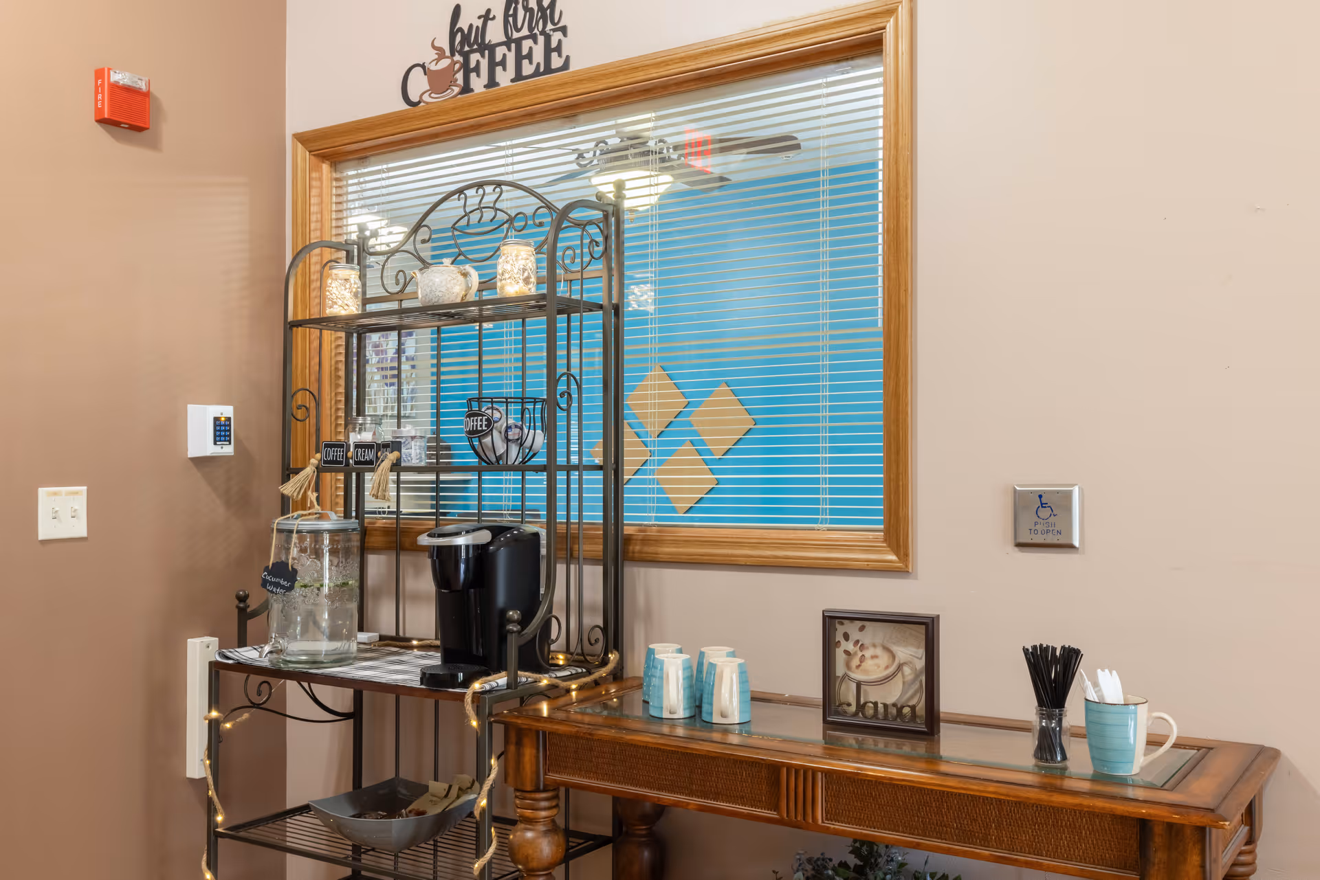 A coffee station with a coffee maker, glass dispensers, cups and utensils on a metal rack and wooden table beneath a window with blinds.