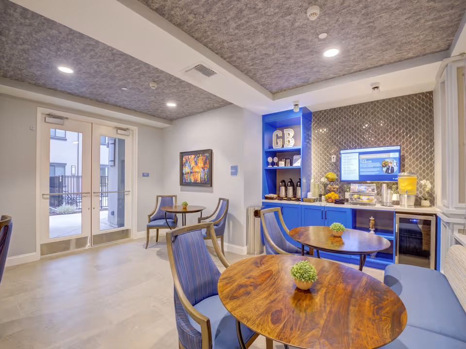 Bright common dining area with round wooden tables, blue chairs, a beverage station and double glass doors to the outside.