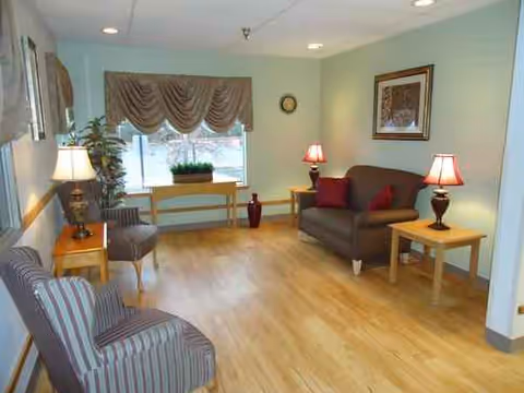 A cozy senior living lounge with sofa, chairs, lamps, and a window view.