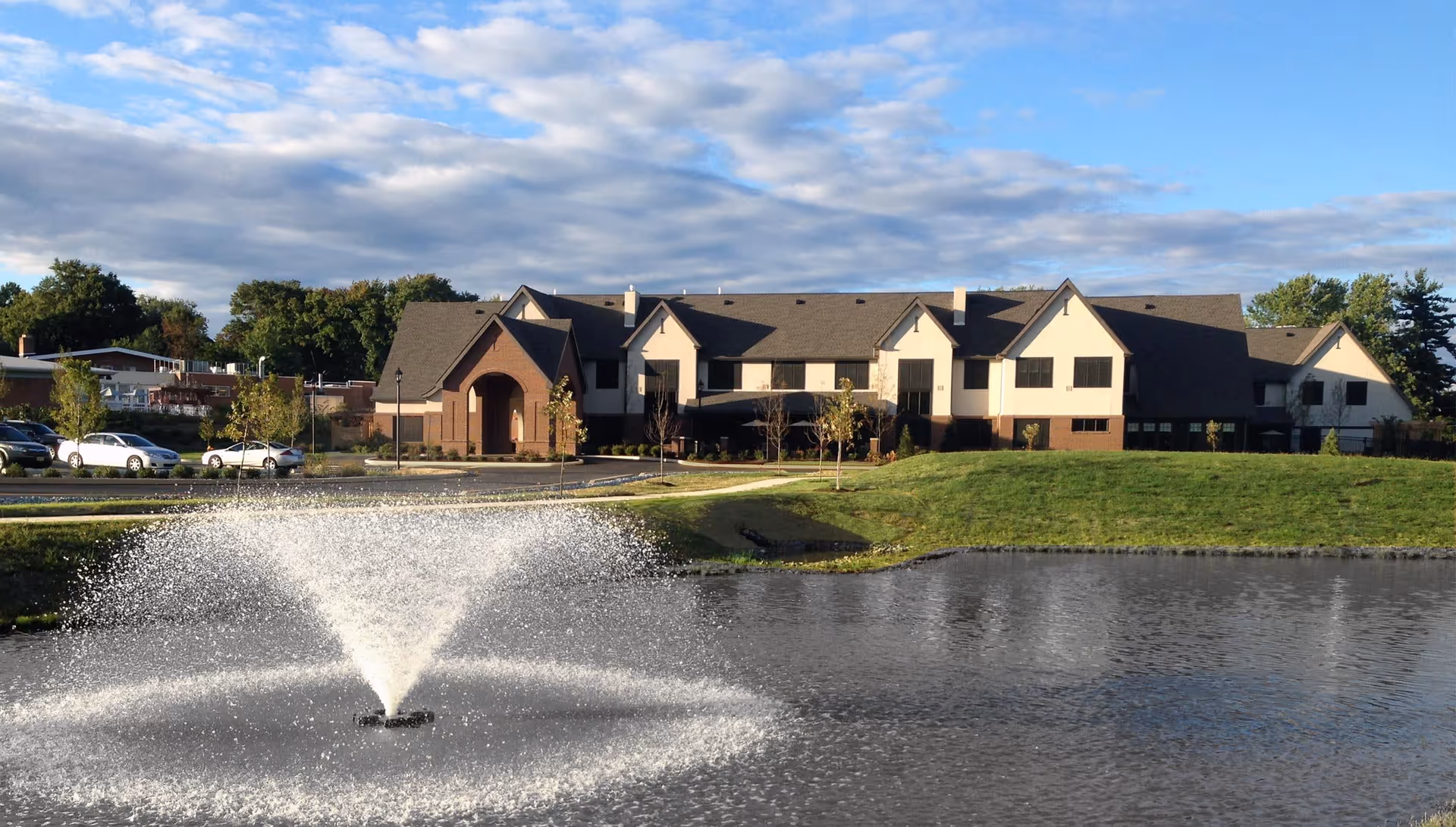 Two-story assisted living building seen across a pond with a central fountain under a partly cloudy sky.