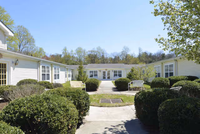 Outdoor courtyard area of a senior living facility named ALC, featuring a paved walkway surrounded by neatly trimmed bushes and green grass, with white benches and single-story white buildings with multiple windows under a clear blue sky.