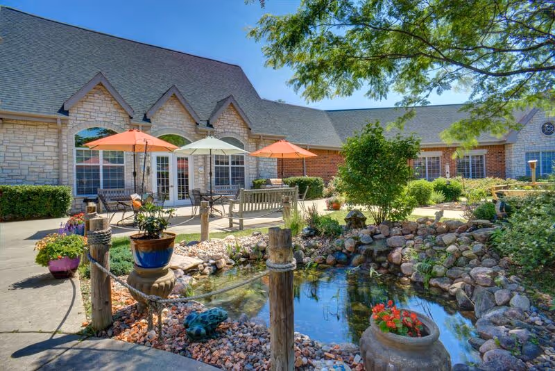 Outdoor garden area at Newcastle Place featuring a small pond surrounded by rocks and plants, wooden posts with rope fencing, potted flowers, benches, and patio tables with colorful umbrellas in front of a stone and brick building under a clear blue sky.