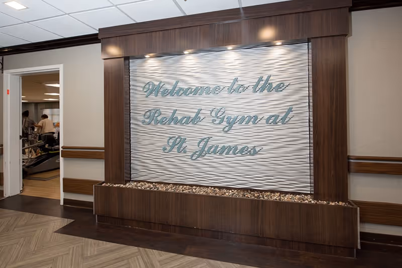 Interior view of a rehabilitation gym entrance at St. James Rehabilitation & Healthcare Center. A decorative wall panel with the text 'Welcome to the Rehab Gym at St. James' is prominently displayed, framed by dark wood. To the left, an open doorway reveals part of the gym area with exercise equipment and two people inside.