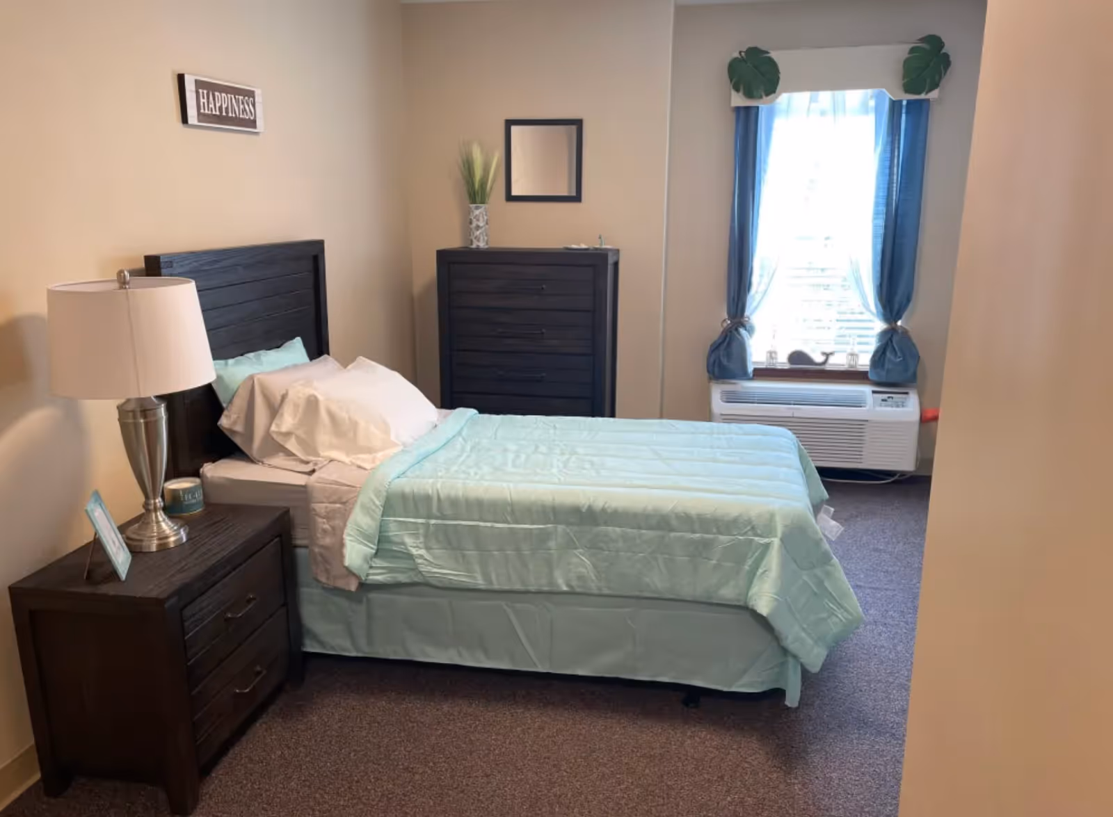 A tidy bedroom with a twin bed dressed in light teal bedding, a dark wood nightstand and dresser, a lamp, and a window with blue curtains and an air-conditioning unit.