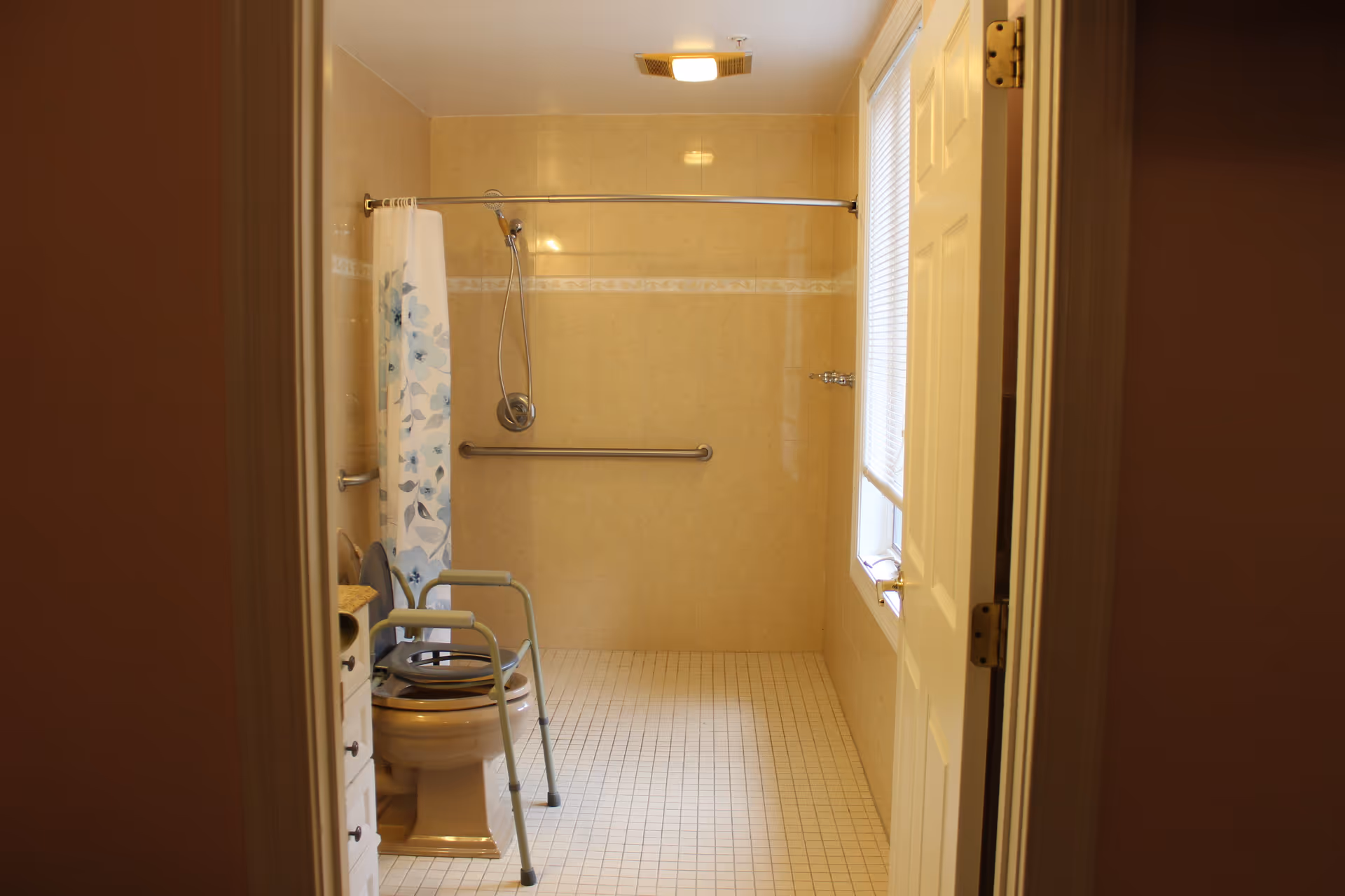 Accessible bathroom with a walk-in shower, grab bar, toilet with a raised commode frame and a floral shower curtain.