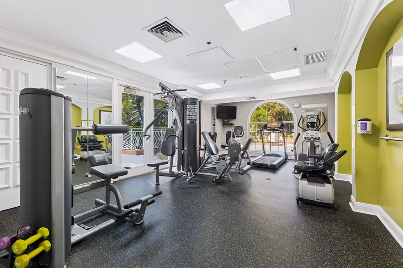 A well-lit fitness room with various exercise equipment including a multi-station gym machine, stationary bikes, and a treadmill. The room has large windows and a glass door leading to an outdoor area with greenery visible outside. The walls are painted in white and green, and there is a wall-mounted TV in the corner.