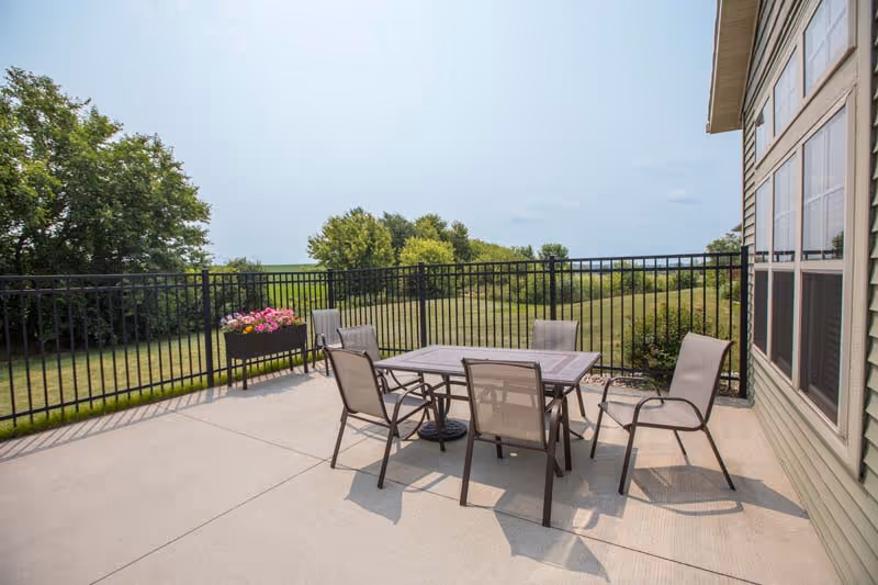 Patio with a table and six chairs on a concrete terrace beside a building, overlooking a grassy yard and trees behind a black metal fence.