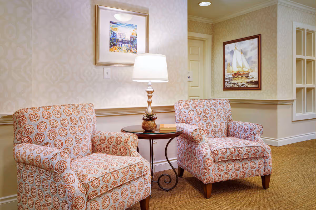 Two patterned armchairs with circular designs are placed on either side of a small round table with a lamp and a small plant on it. The room has beige wallpaper with a subtle pattern, a carpeted floor, and framed artwork on the walls, including a painting of a sailboat and a colorful street scene.