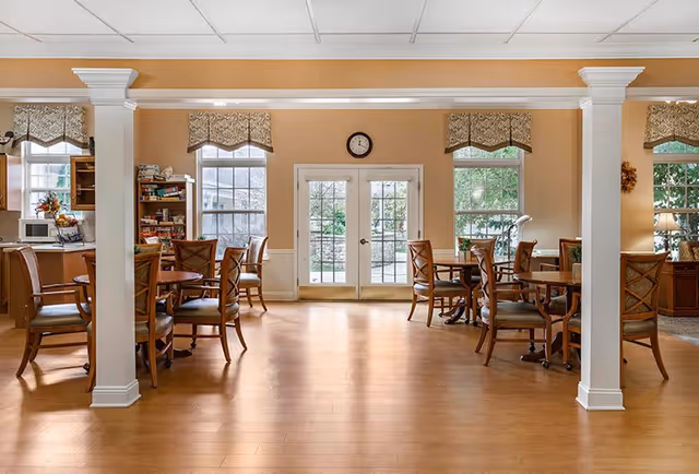 A bright dining area with wooden floors, several round tables surrounded by wooden chairs with cushions, beige walls, and large windows with patterned valances. French doors lead outside, and a clock is mounted above the doors. The room has white columns and a cozy, inviting atmosphere.