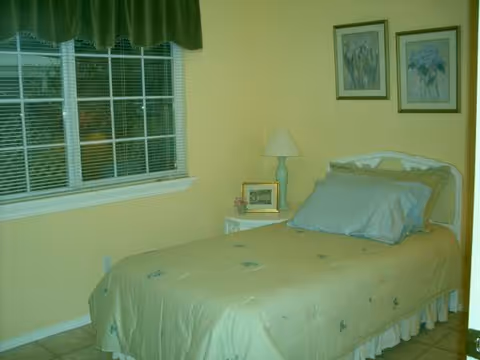 Single bedroom with bed, nightstand, lamp, window, and framed pictures in pale yellow room.