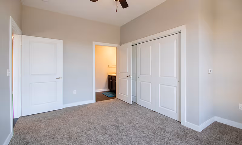 Empty bedroom with beige carpet, light gray walls, white doors including a closet with sliding doors, and an open door leading to a bathroom with a dark vanity and a blue bath mat.
