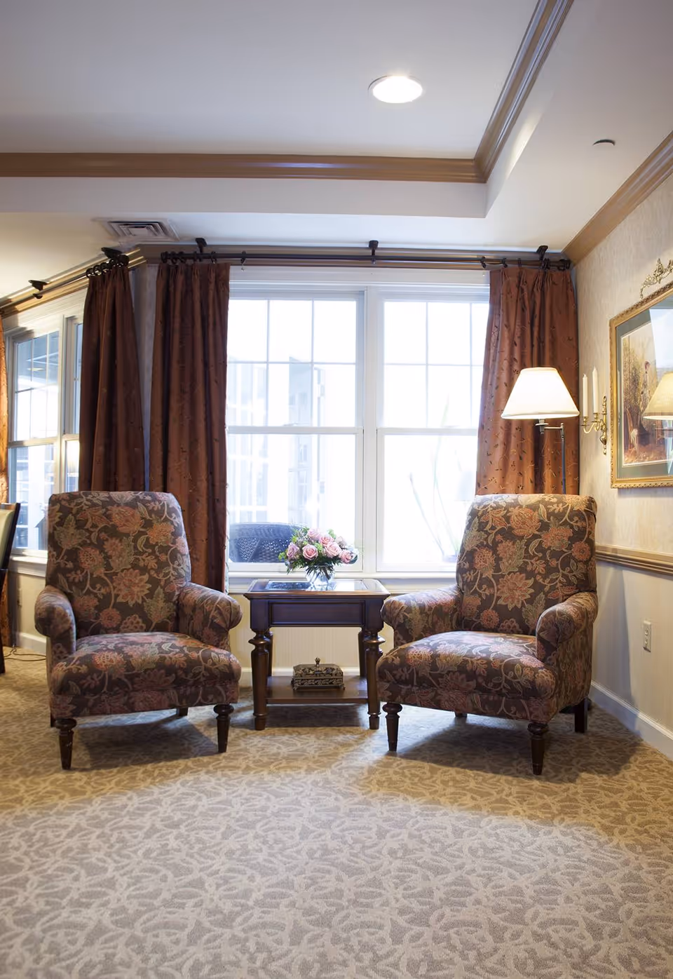 Two floral-patterned armchairs with wooden legs are positioned on either side of a small wooden table with a glass vase of pink flowers. Behind them is a large window with brown curtains, and a floor lamp is next to the right chair. The room has beige patterned carpet and light-colored walls with a framed painting.