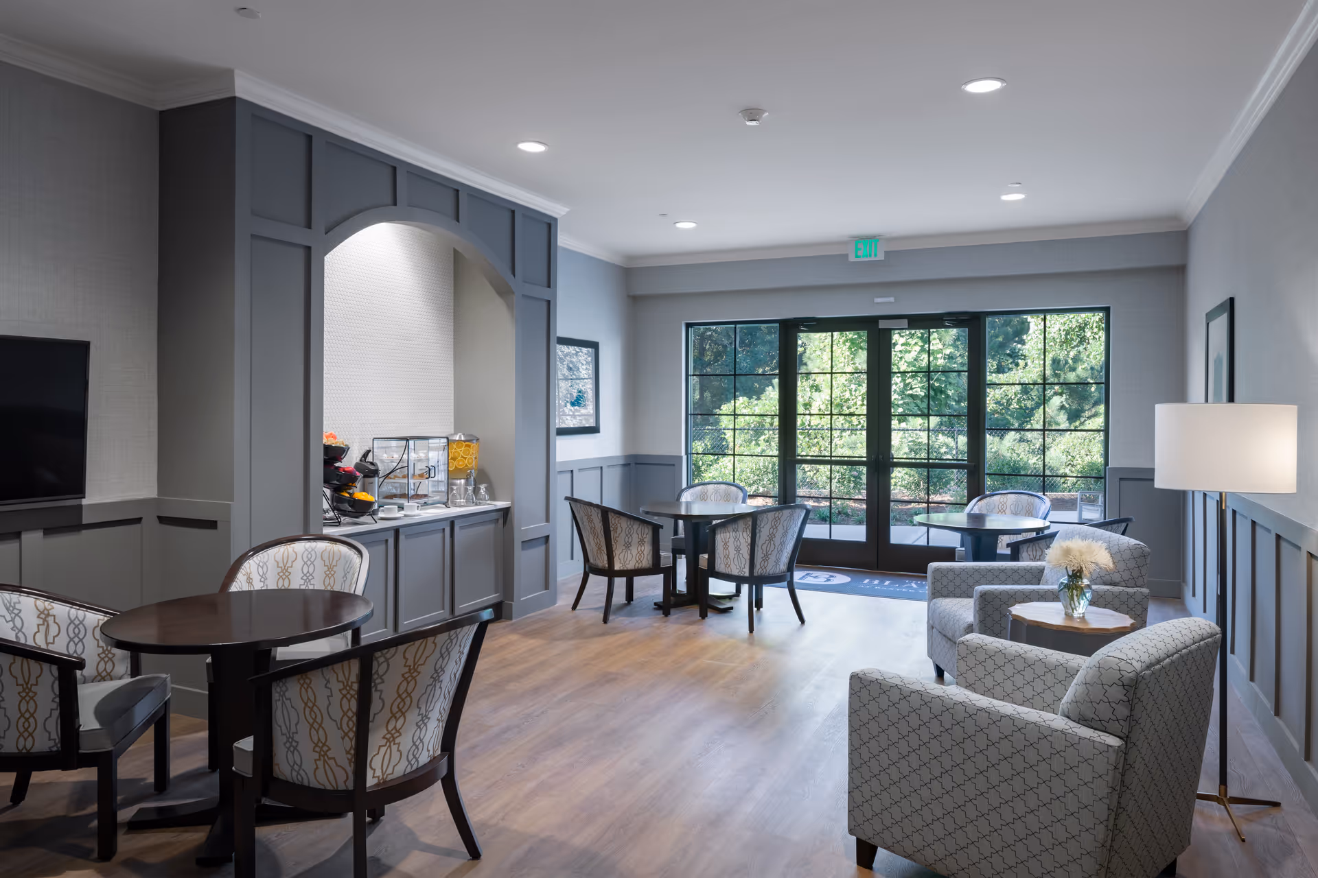 A bright and modern common area with several small round tables and upholstered chairs arranged for seating. There is a built-in counter with a beverage dispenser and snacks under an archway on the left side. Large windows and glass doors at the back provide a view of greenery outside. The room features wood flooring, gray wainscoting, and soft lighting from ceiling fixtures and a floor lamp.