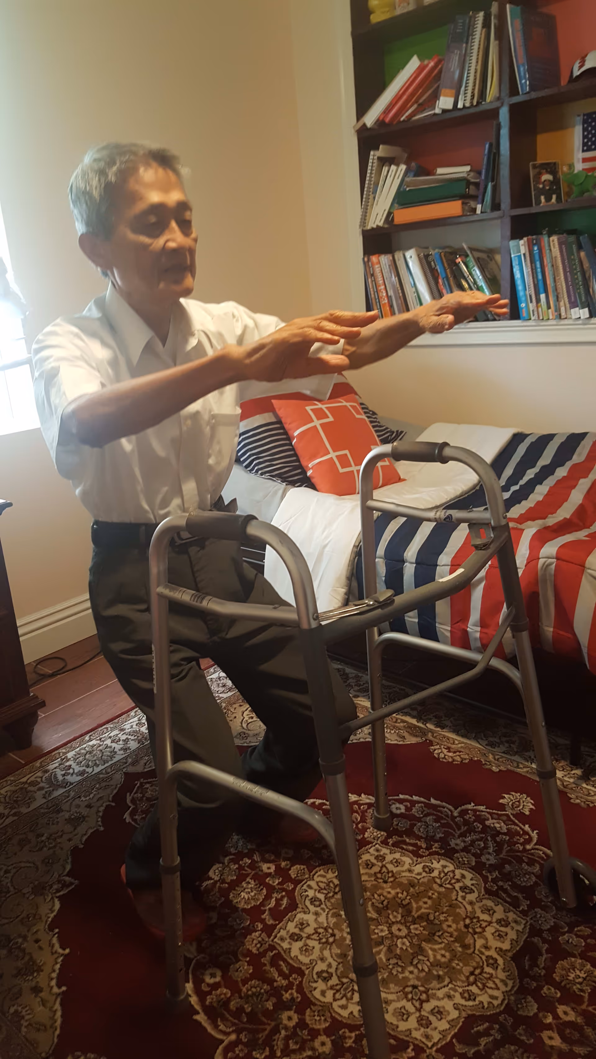 An elderly man using a walker in a bedroom with a bed covered in a striped red, white, and blue blanket. There is a bookshelf filled with books and some decorative items behind the bed. The room has a patterned rug on the floor and a window letting in natural light.