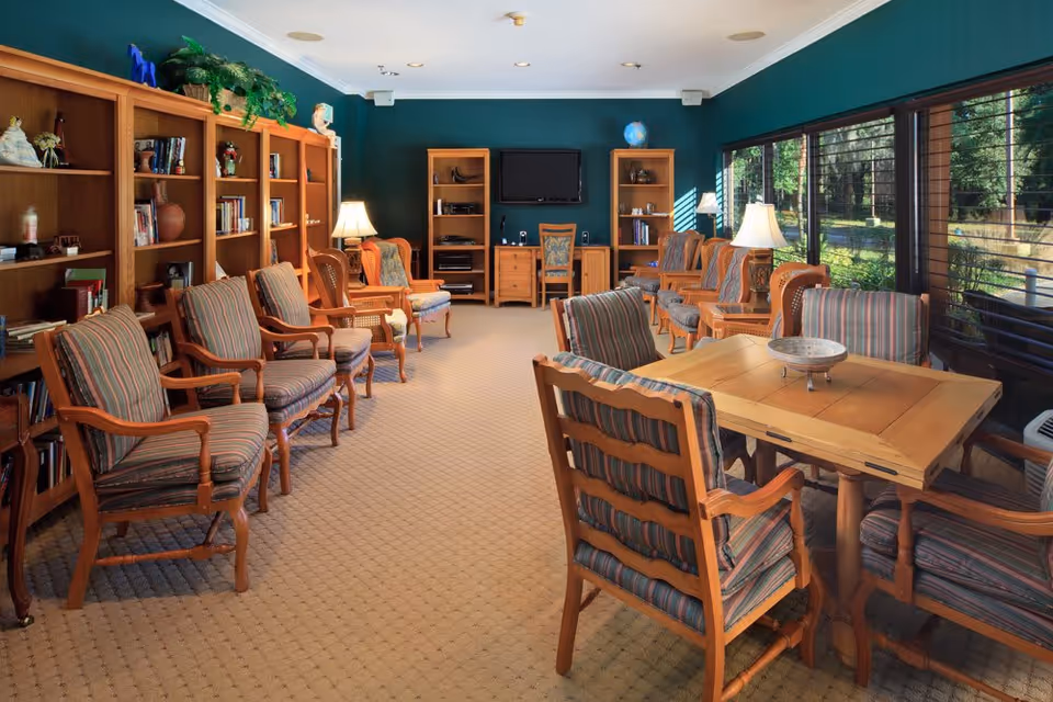 A cozy living room with multiple wooden chairs upholstered in striped fabric arranged around a wooden table and along the walls. The room features wooden bookshelves filled with books and decorative items, a flat-screen TV mounted on the wall, and large windows with blinds letting in natural light and offering a view of greenery outside. Several table lamps provide warm lighting.