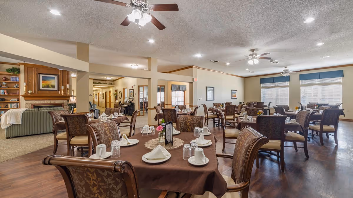 A spacious dining room in a senior living facility with multiple tables covered in brown tablecloths, each set with white plates, cups, napkins, and glasses. The room features ceiling fans with lights, large windows with blue valances, and a cozy seating area with a fireplace and bookshelves in the background.