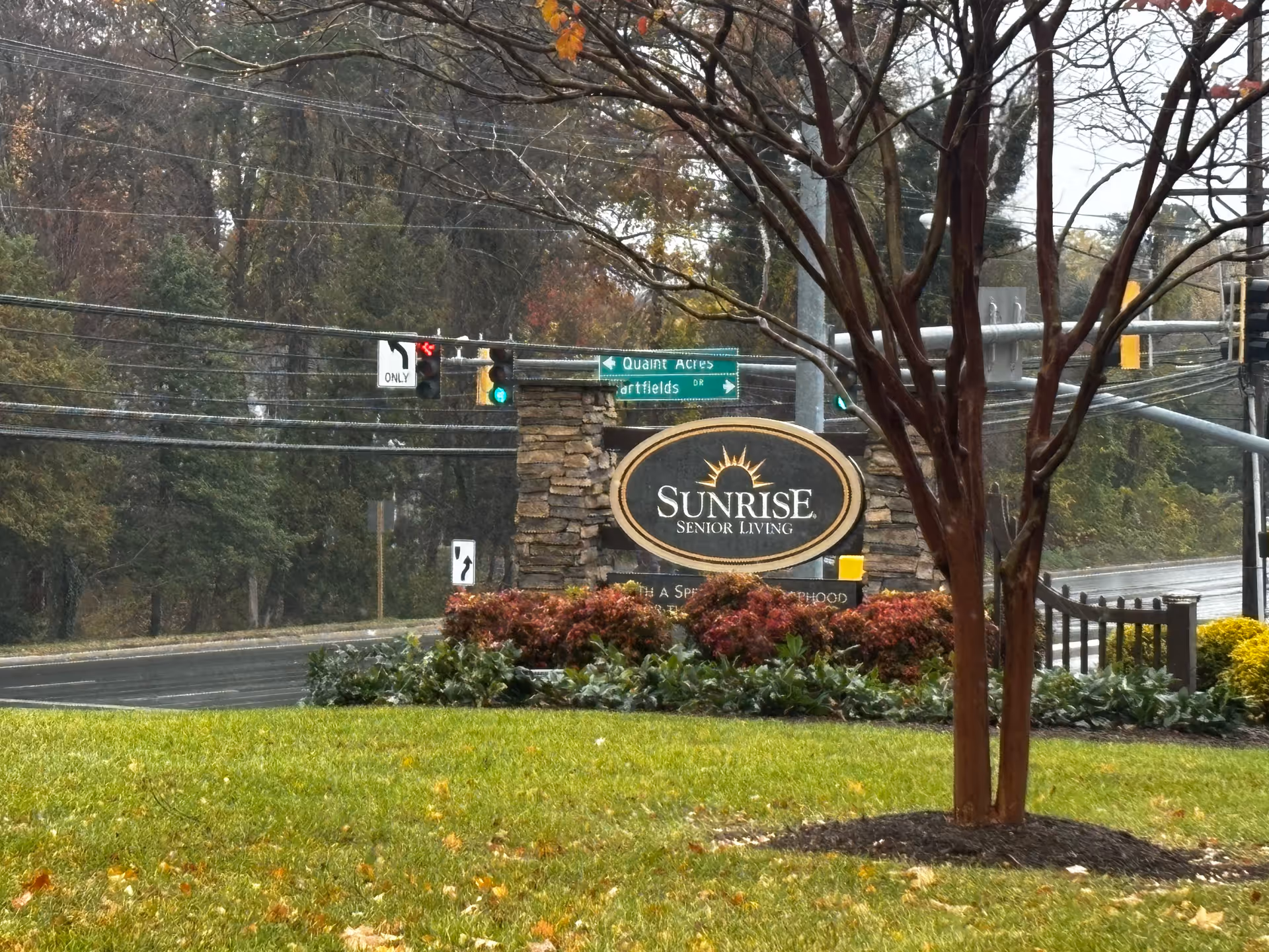 Outdoor view of the entrance sign for Sunrise Senior Living, surrounded by landscaping with green grass, bushes, and a tree in the foreground. Traffic lights and road signs are visible in the background along a street lined with trees.