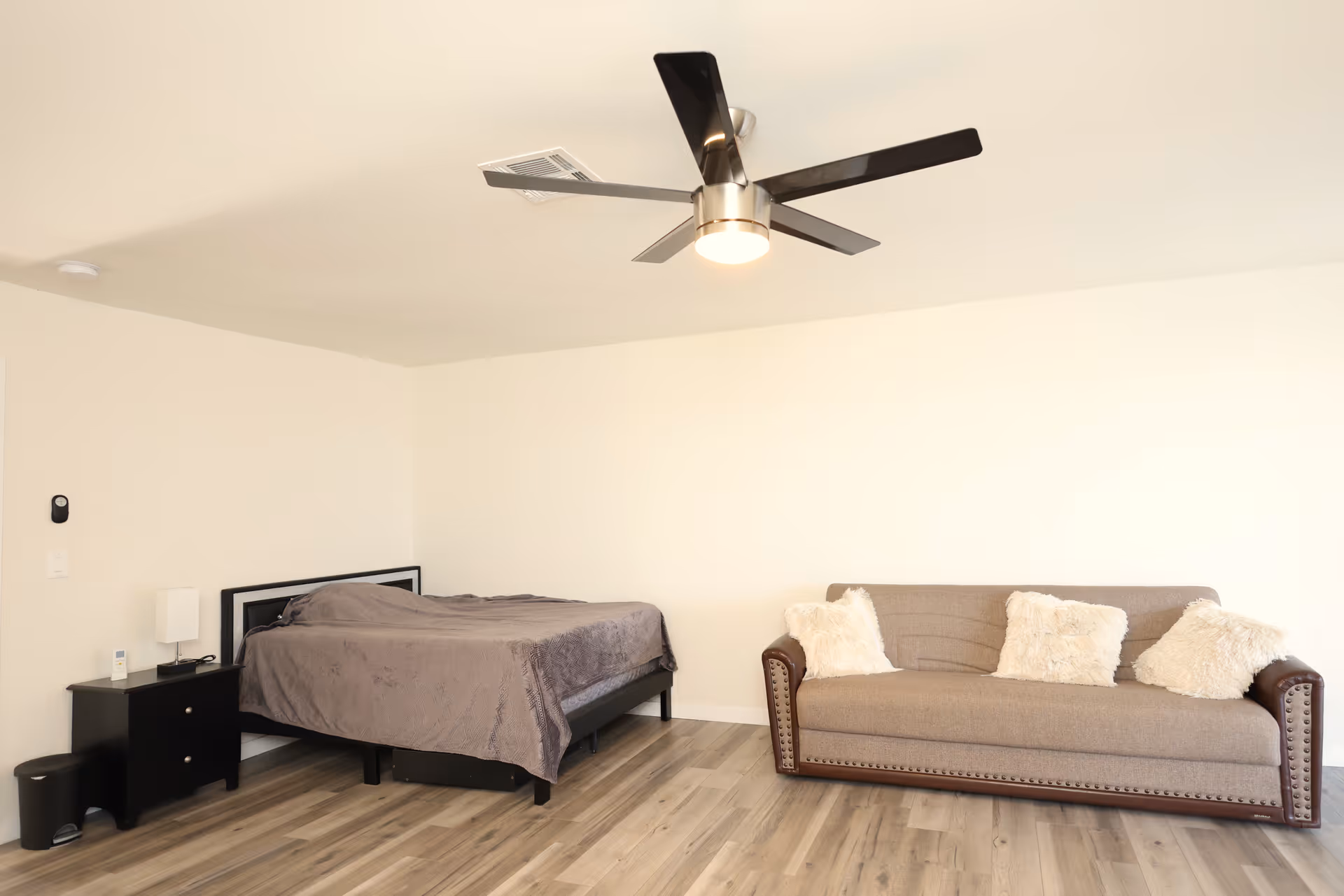 A simple, clean room with a bed covered in a gray blanket, a black nightstand with a lamp and small items, a beige sofa with three fluffy white pillows, and a modern ceiling fan with five blades. The room has light-colored walls and wood flooring.