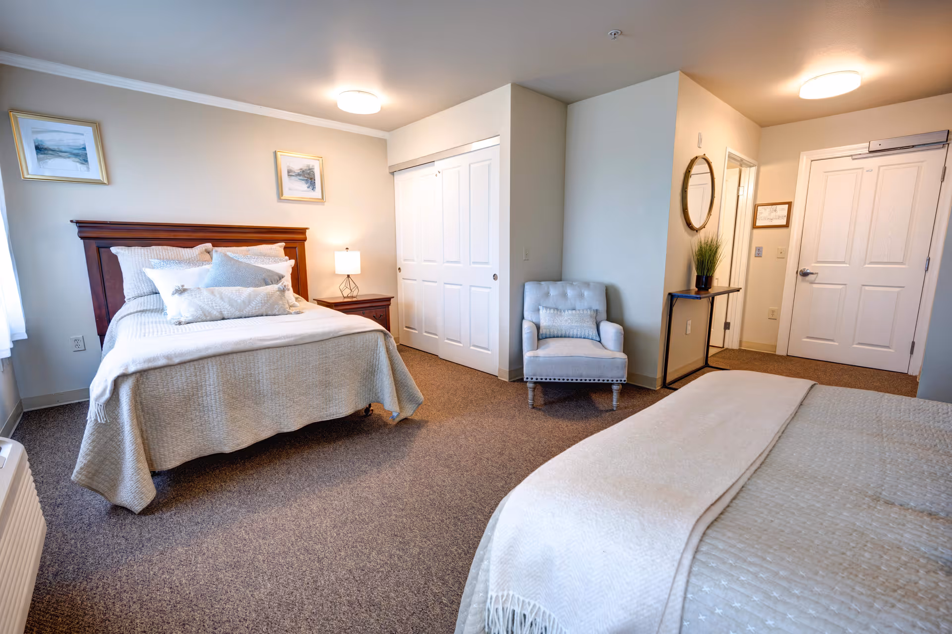 A bright and clean bedroom in a senior living facility with two beds covered in beige bedding. There is a wooden headboard on one bed, a nightstand with a lamp, two framed pictures on the wall, a light blue armchair with a pillow, a small table with a plant and a round mirror above it, and a closed white closet door. The room has beige carpet and white walls.