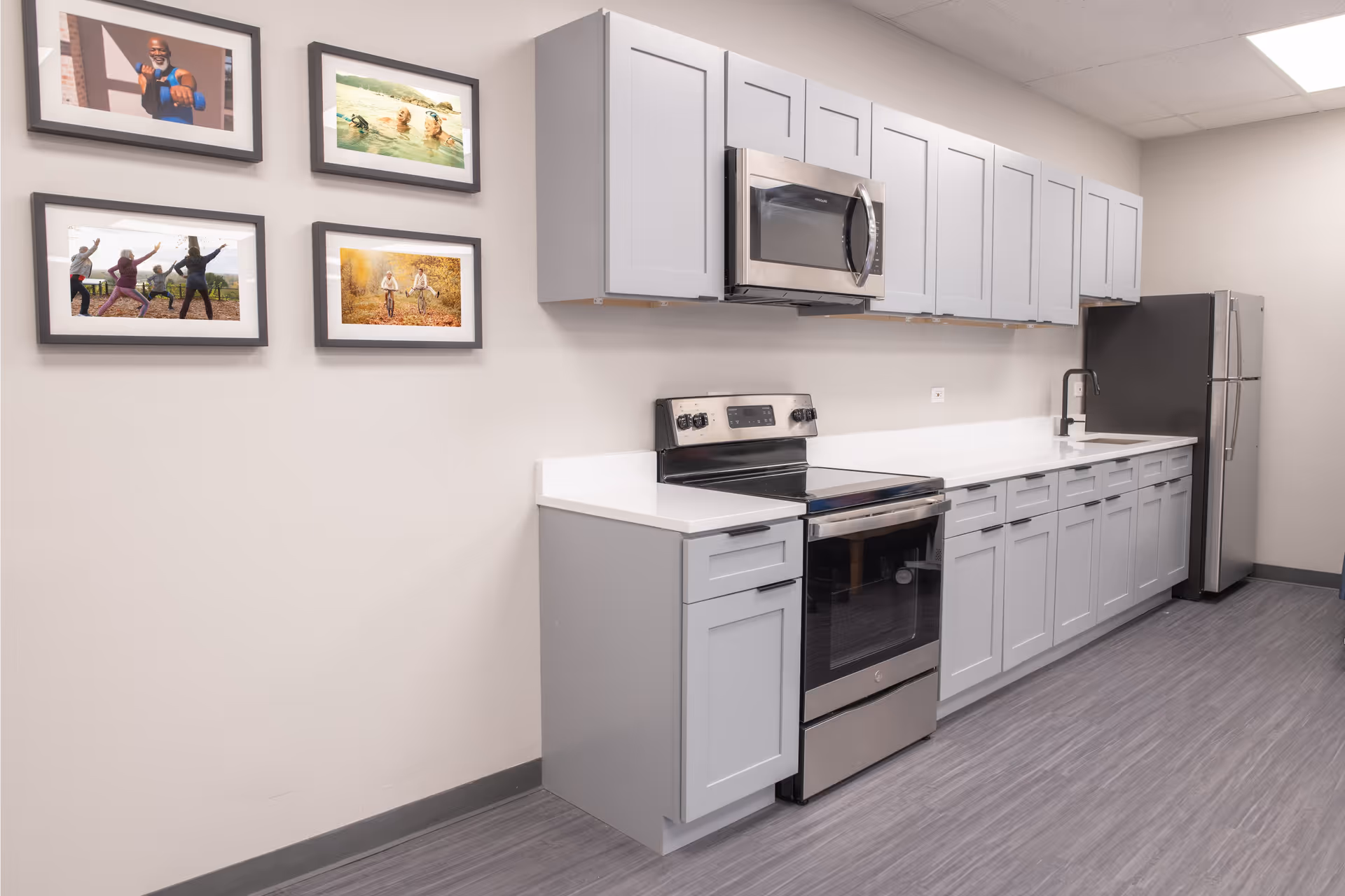 Modern kitchen with light gray cabinets, stainless steel appliances including a stove, microwave, and refrigerator, white countertops, and four framed photos on the adjacent wall.