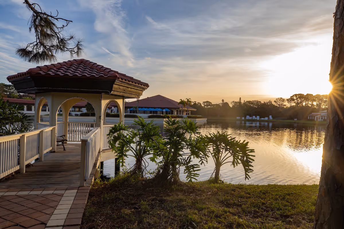 A wooden gazebo with a red tiled roof extends over a calm lake at sunset, surrounded by greenery and trees, with buildings visible in the background under a partly cloudy sky.