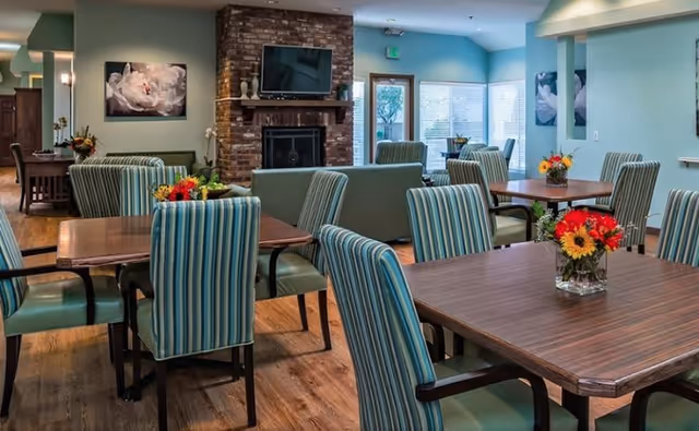 A bright and inviting dining area in a senior living facility with wooden tables and striped upholstered chairs. Each table has a small vase with colorful flowers. There is a brick fireplace with a TV mounted above it, and large windows letting in natural light. The walls are painted in soft green and blue tones, and there are floral paintings on the walls.