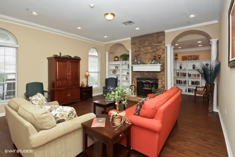 A cozy living room with a beige sofa and a red sofa facing a stone fireplace. The room features wooden flooring, built-in white shelves on either side of the fireplace, and arched doorways. There are two windows with blinds, a wooden cabinet, a green armchair, and various decorative plants and items.